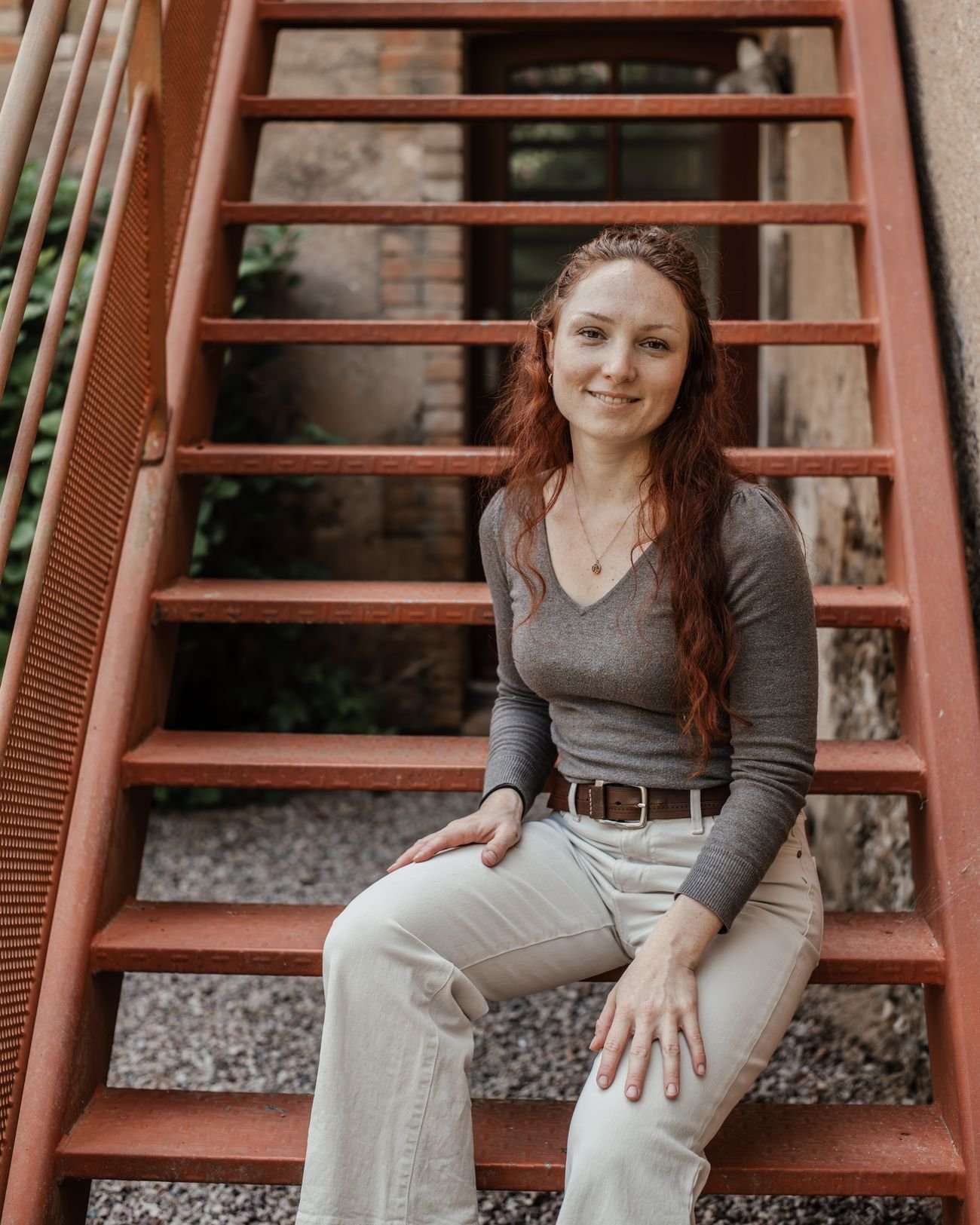 Portrait de Laura Bridier, souriante, assise sur un escalier extérieur en bois rouge.